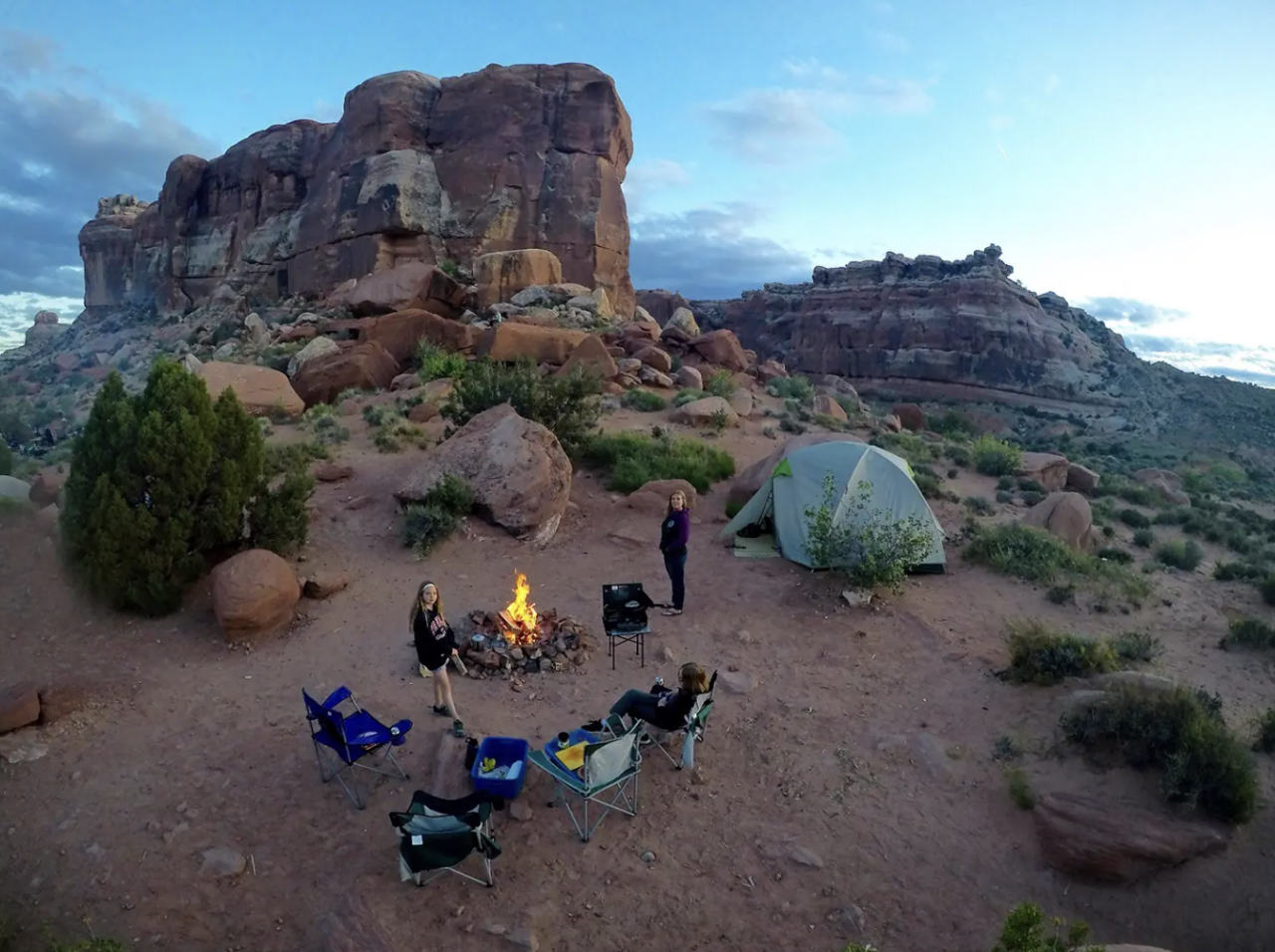 Camping in the desert with a tent, fire, and campers relaxing in camping chairs.